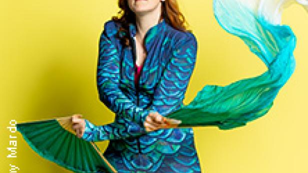 Eine Frau in einem blaugrünen Kostüm posiert mit einem Fächer und einem wehenden Tuch vor einem gelben Hintergrund.