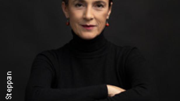 Eine Frau mit dunklen Haaren und einem schwarzen Rollkragenpullover sitzt auf einem Stuhl.