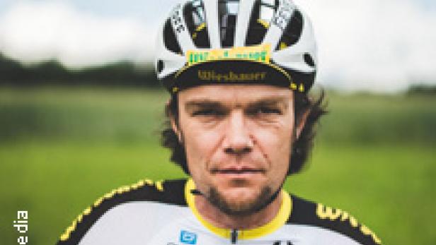 Ein Radfahrer in Sportkleidung und Helm steht vor einer grünen Landschaft.