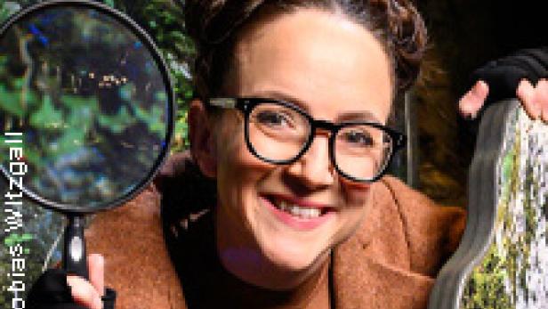 Eine lächelnde Frau mit Brille und Dutt hält eine Lupe vor einem grünen Hintergrund.