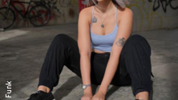 Eine junge Frau mit hellblondem Haar sitzt vor einer Graffitiwand.