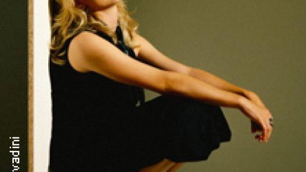 Eine Frau in einem schwarzen Kleid und weißen Stiefeln kauert vor einer Wand.