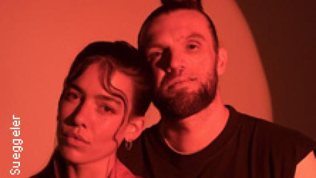 Ein Mann mit Dutt und eine Frau mit Blazer posieren vor einem roten Hintergrund.