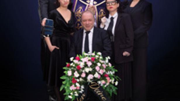 Eine Gruppe von Personen in Trauerkleidung steht um einen Kranz mit der Aufschrift „Ruhe in Frieden, Deine Mutter Mitarbeiter Team“.