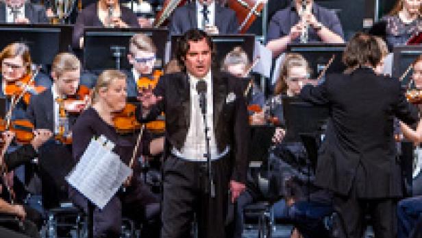 Ein Sänger steht vor einem Orchester und singt in ein Mikrofon.