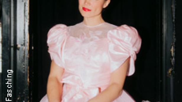 Eine Frau in einem rosa Kleid sitzt vor einem dunklen Hintergrund.