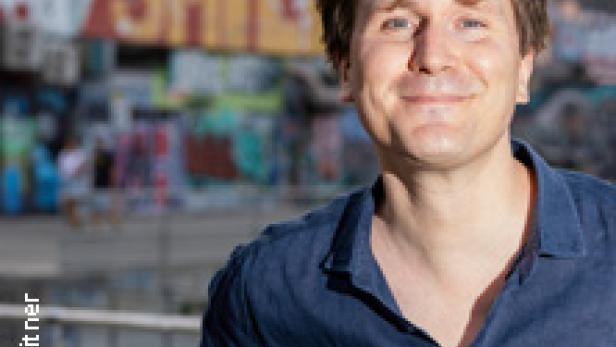 Ein Mann mit blauem Hemd lächelt vor einer Wand mit Graffiti.