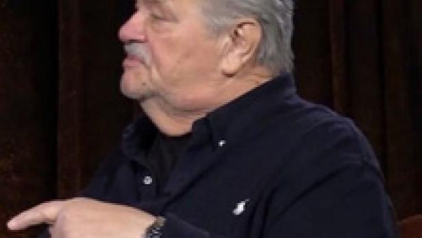 Ein Mann mit grauem Haar und Schnurrbart zeigt mit dem Finger nach links.