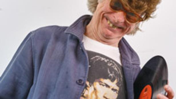 Ein Mann mit Perücke und Sonnenbrille lächelt und hält eine Schallplatte.