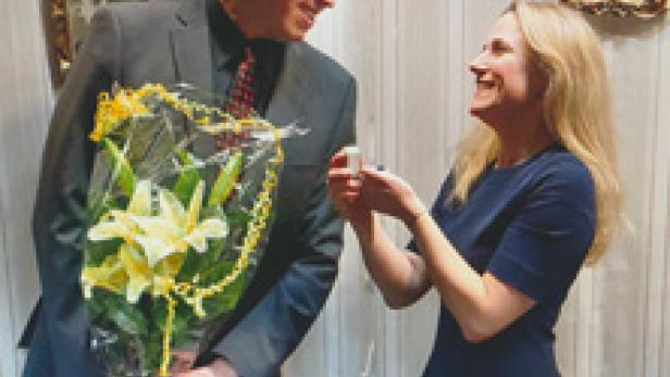 Ein Mann mit Blumenstrauß hält um die Hand einer lachenden Frau an.