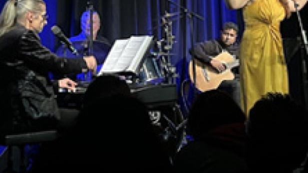 Eine Sängerin tritt mit ihrer Band auf einer Bühne vor einem dunklen Vorhang auf.