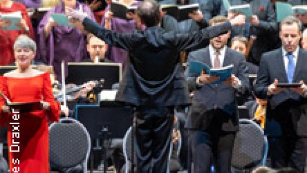 Ein Dirigent steht vor einem Chor und einem Orchester während einer Aufführung.