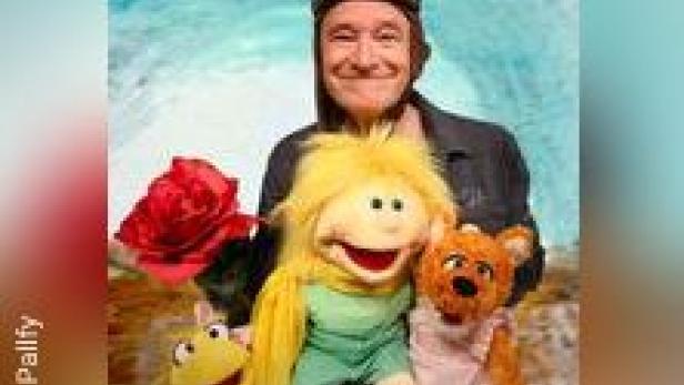 Ein Mann mit Fliegerbrille posiert mit vier bunten Handpuppen und einer roten Rose vor einem hellblauen Hintergrund.