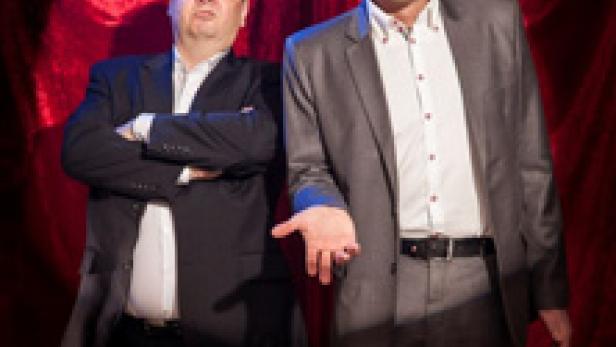 Zwei Männer in Anzügen stehen vor einem roten Vorhang.