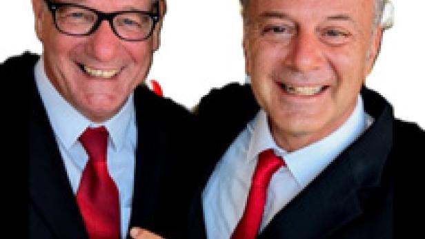 Zwei Männer in Anzügen mit roten Krawatten lächeln in die Kamera.