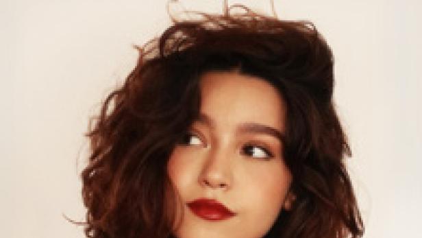 Porträt einer jungen Frau mit dunklen Haaren und rotem Lippenstift vor einem Ankündigungsplakat für Mari Froes in Wien.