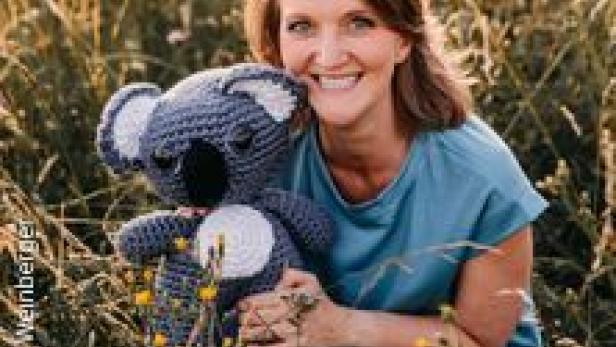 Eine Frau hockt in einem Feld und hält einen gehäkelten Koala-Bären.