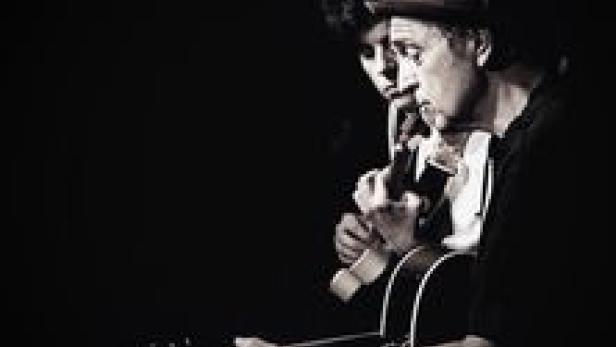 Ein Mann mit Hut spielt Gitarre, während eine Frau ihm aufmerksam zuhört.