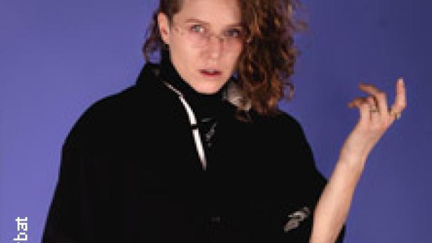 Eine Frau mit lockigem Haar und schwarzer Jacke posiert vor einem blauen Hintergrund.