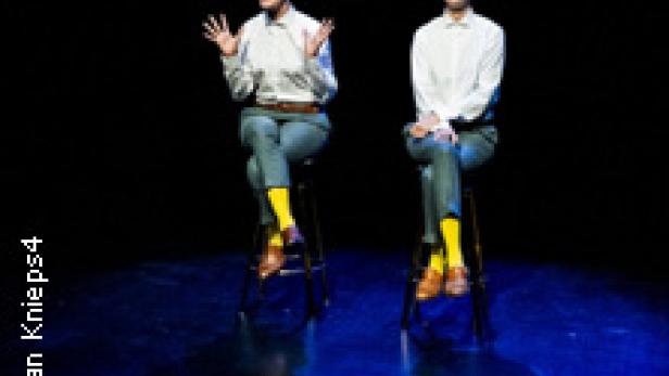 Zwei Personen sitzen auf Stühlen und scheinen auf einer Bühne zu performen.