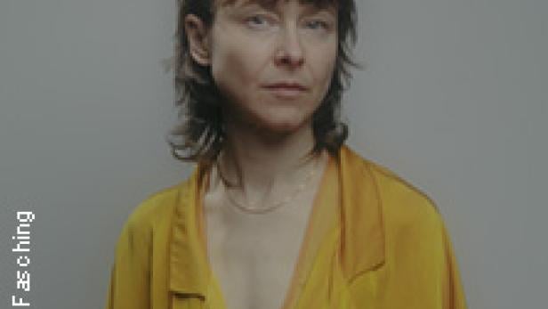 Porträt einer Frau mit blonden Haaren und gelber Bluse vor einem grauen Hintergrund.