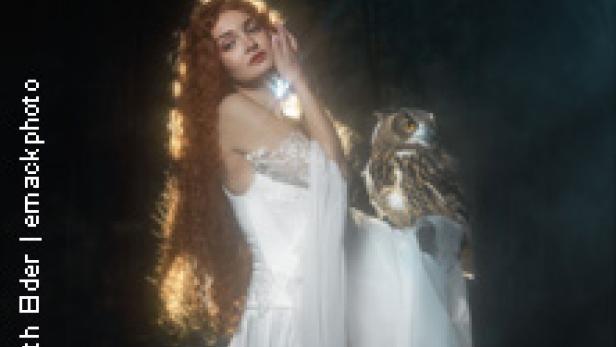 Eine Frau in einem weißen Kleid mit einer Eule auf der Schulter vor dunklem Hintergrund.