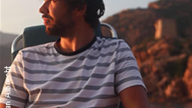 Ein Mann mit Bart und Kappe sitzt am Strand und schaut in die Ferne.