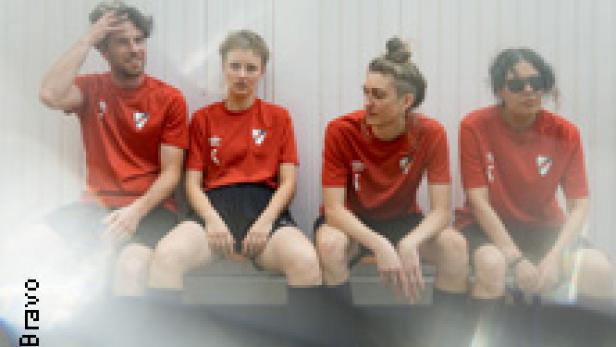Vier Personen in roten Fußballtrikots sitzen auf einer Bank vor einer weißen Wand.