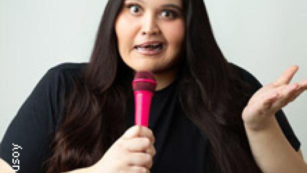 Eine Frau mit langen, dunklen Haaren hält ein pinkfarbenes Mikrofon und spricht.