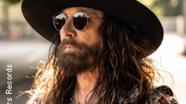 Ein Mann mit Bart, Hut und Sonnenbrille posiert für ein Foto.