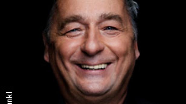 Ein lächelnder Mann mit kurzen, grauen Haaren vor einem schwarzen Hintergrund.