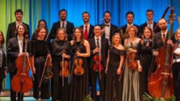 Ein Orchester steht auf einer Bühne vor einem blauen Vorhang.