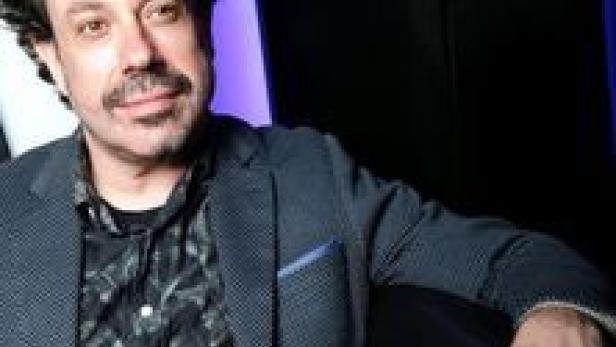 Ein Mann mit dunklen, lockigen Haaren und Bart trägt ein dunkles Sakko.