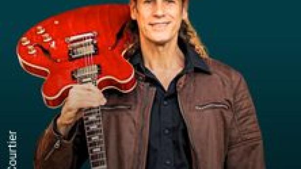 Ein Mann mit Hut und Gitarre posiert vor einem türkisfarbenen Hintergrund.