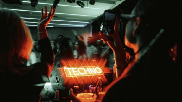 Blick vom DJ-Pult auf den Innenraum. Menschen haben die Hände in der Höhe und tanzen, eine Person fotografiert.. Auf einem Leuchtschild steht Techno.
