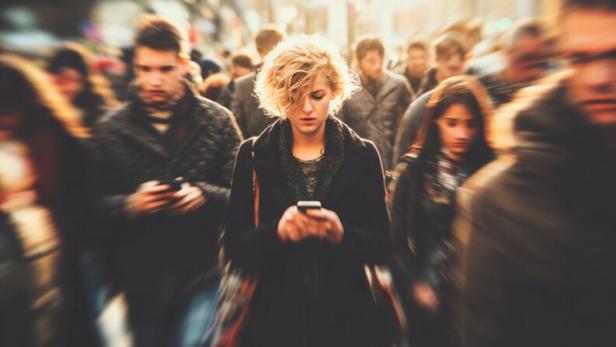 Eine junge Frau mit lockigen Haaren steht inmitten einer Menschenmenge und blickt auf ihr Smartphone.