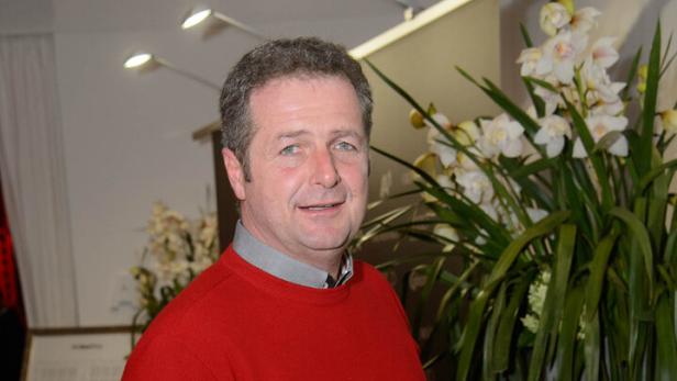 Norbert Rier mit rotem Pullover steht neben einer hohen Pflanze mit weißen Blüten.