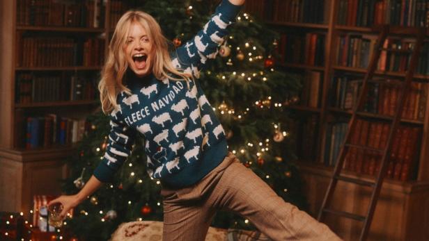 Eine blonde Frau im Weihnachtspullover mit Schafen jubelt vor einem Weihnachtsbaum und Bücherregalen.