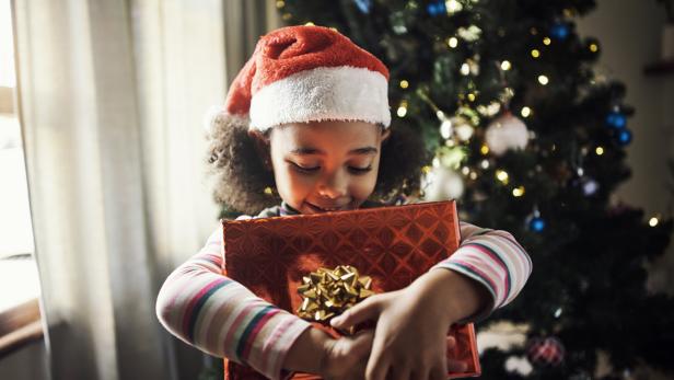 Ein Kind mit Weihnachtsmütze umarmt ein Geschenk vor einem festlich geschmückten Weihnachtsbaum.