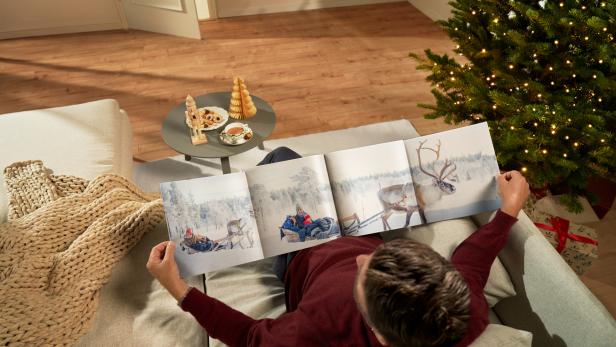 Mann betrachtet auf dem Sofa ein Fotobuch mit Wintermotiven neben einem geschmückten Weihnachtsbaum.