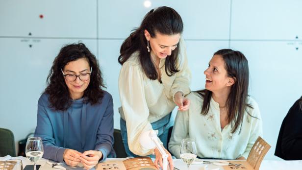 Drei Frauen sitzen an einem Tisch und arbeiten gemeinsam an Schmuckstücken, während eine lächelnd Anweisungen gibt.