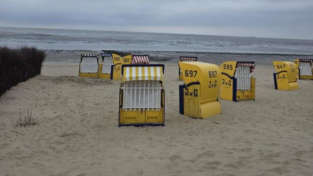 Gelbe Strandkörbe auf leerem Sandstrand vor grauer See und bewölktem Himmel.