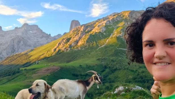 Eine Frau macht ein Selfie mit einem Hund und einer Ziege vor einer sonnigen Bergkulisse.