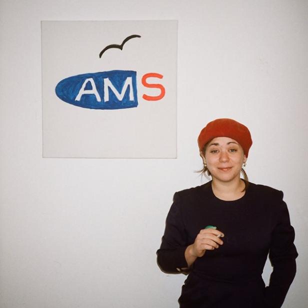 Stefanie Sargnagel steht vor einem Bild mit der Aufschrift „AMS“.