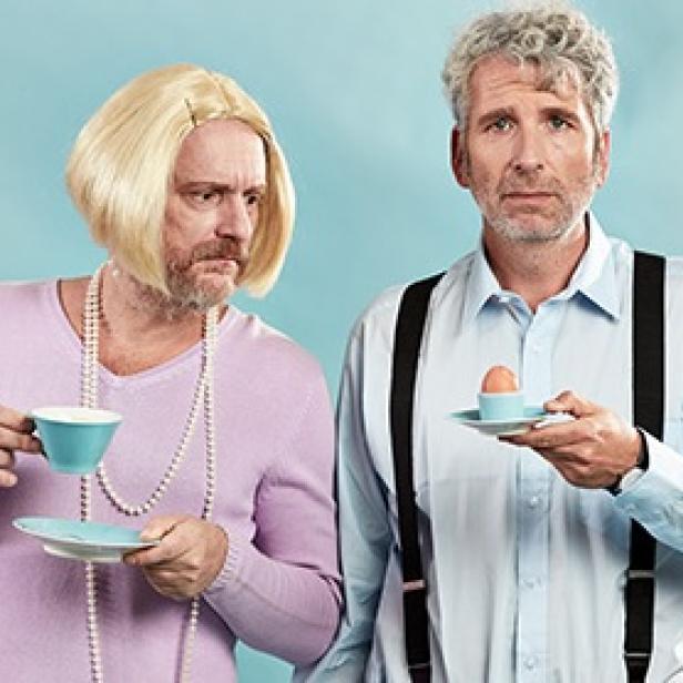 Zwei Männer, einer mit Perücke und Teetasse, der andere mit Hosenträgern und einem Frühstücksei.