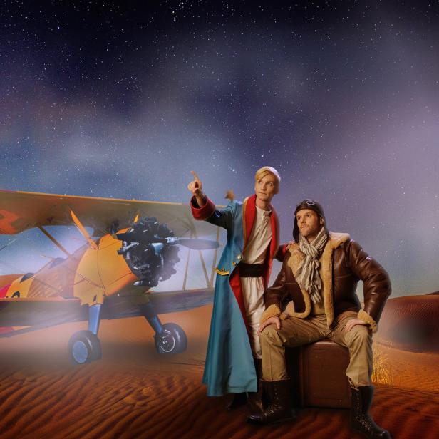 Der kleine Prinz und ein Pilot stehen vor einem gelben Doppeldecker in der Wüste.