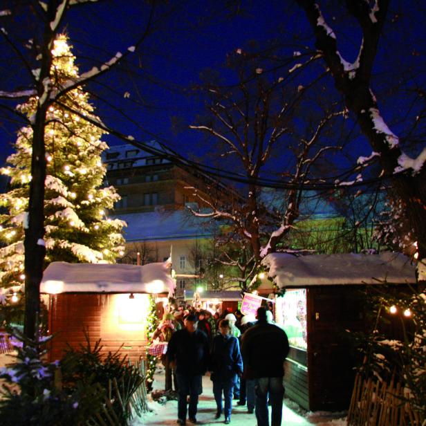 Ein belebter Weihnachtsmarkt mit schneebedeckten Hütten und einem hell erleuchteten Weihnachtsbaum.