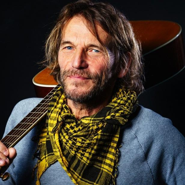 Hans Söllner mit Gitarre und gelb-schwarzem Tuch vor dunklem Hintergrund.