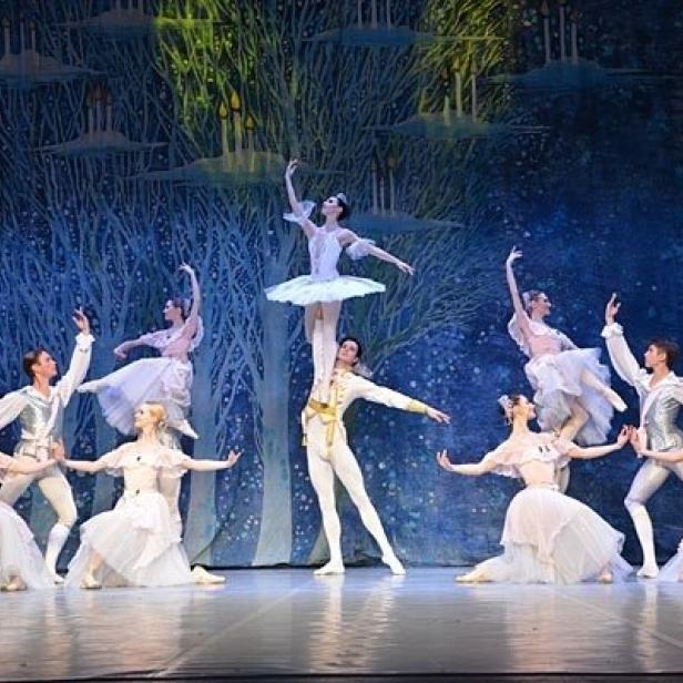 Eine Ballettaufführung mit Tänzern in weißen Kostümen vor einem Bühnenbild mit Bäumen.