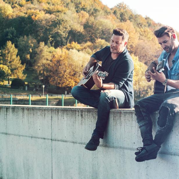 Zwei Männer sitzen auf einer Mauer und spielen Gitarre vor einer herbstlichen Waldkulisse.